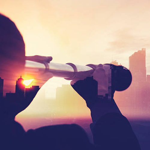 Business man in suit with cityscape montage. The man is unrecognizable and you cannot see his face. He is superimposed onto a city skyline at sunset. He is holding a telescope looking into the city. Success, vision concept with copy space.
